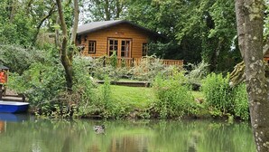 A wooden cabin with a dock and boats, surrounded by lush greenery and a calm lake.