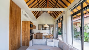 A modern living room with a large sectional sofa, a wooden coffee table, and a kitchen area with wooden cabinets and a hanging pendant light. - Gaze Villa by Betterplace (Canggu)