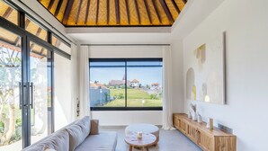 A modern living room with a large window, a sofa, a round table, and a wooden console. - Gaze Villa by Betterplace (Canggu)