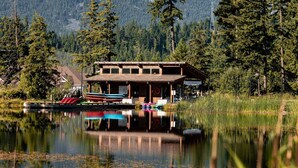 Un chalet avec une façade en bois, un quai avec des canoës et une chaîne de montagnes en arrière-plan.
