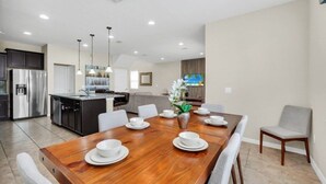 A modern kitchen with stainless steel appliances, a dining area with a wooden table and white chairs, and a living area with a sofa and a TV.