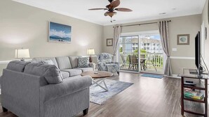 Ein Wohnzimmer mit grauem Sofa, einem Couchtisch, einem Deckenventilator, einer TV-Konsole und Blick ins Freie durch eine Glastür.