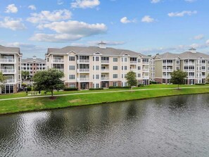 A multi-story residential building with balconies, a grassy area, and a body of water in front. - Magnolia 302 · Golf & Lake Hideaway 3BR (Myrtle Beach)
