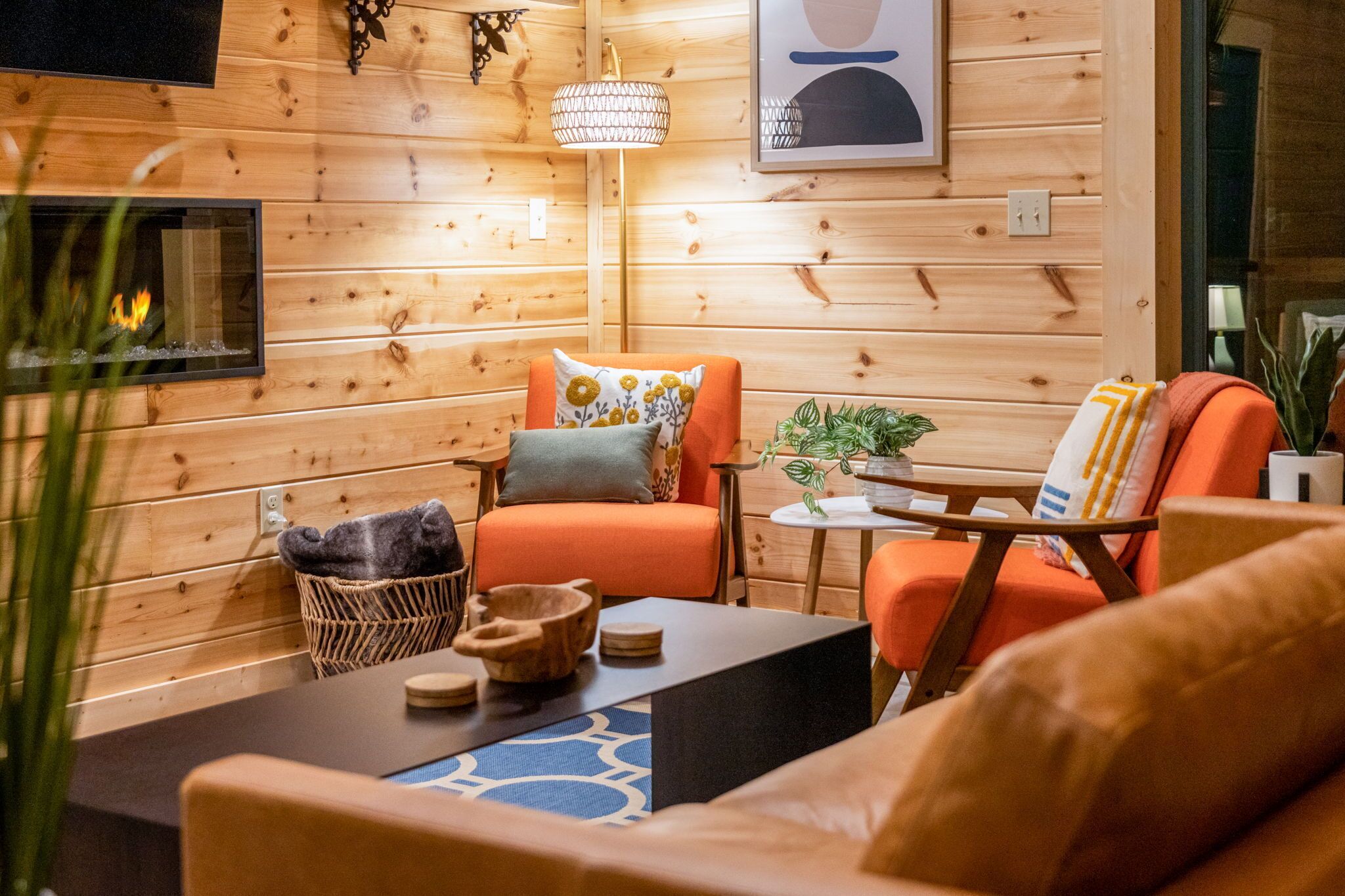 Una habitación acogedora con paredes de madera, una chimenea, sillones naranjas, una mesita y una lámpara colgante.