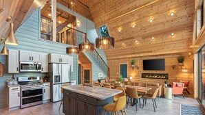A modern kitchen with a central island, a dining area with chairs, a staircase, and a flat-screen TV mounted on the wall. - The Majestic Dragonfly Inn, overlooking Daniel Boone National Forest  (Frenchburg)