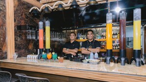 Zona de bar con dos cocteleros, una barra de mármol y una variedad de bebidas coloridas.