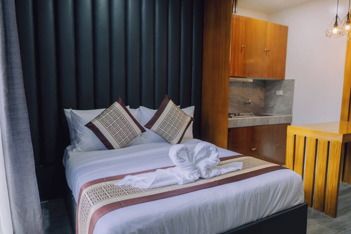 A hotel room with a bed, a wooden wardrobe, a small wooden table, and a modern kitchen area.
