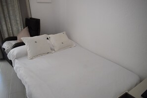 Ein ordentlich bezogenes Bett mit weißer Bettwäsche und zwei Kissen, ein dunkles Kopfholz und eine schlichte Wand mit Lichtschalter.
