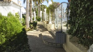 A narrow pathway flanked by lush greenery and a white building with a curved roof.