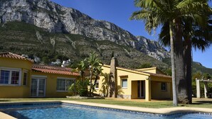 Zona de piscina amb muntanyes al fons, un edifici groc amb teulada vermella i palmeres.