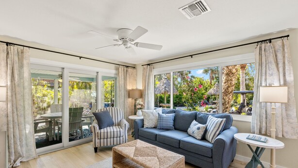 A spacious living room with a grey sofa, striped armchair, and a woven coffee table. Large windows offer a view of a tropical garden. - Driftwood Inn (Sanibel)