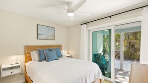 A bedroom with a bed, nightstands, a ceiling fan, a window with a view of greenery, and a sliding glass door. - Driftwood Inn (Sanibel)
