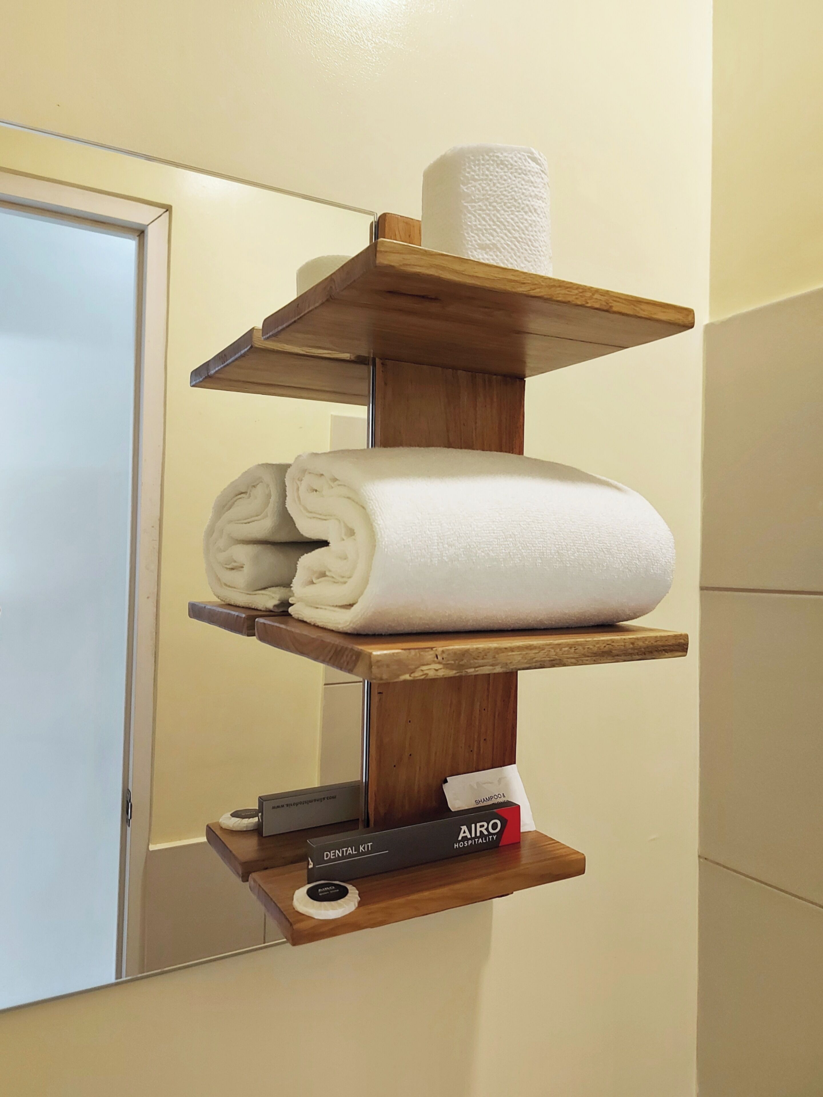 Ein Holzregal mit zusammengerollten Handtüchern und einer Toilettenpapierrolle. Darunter befinden sich ein Dentalset und eine Shampoo-Flasche.