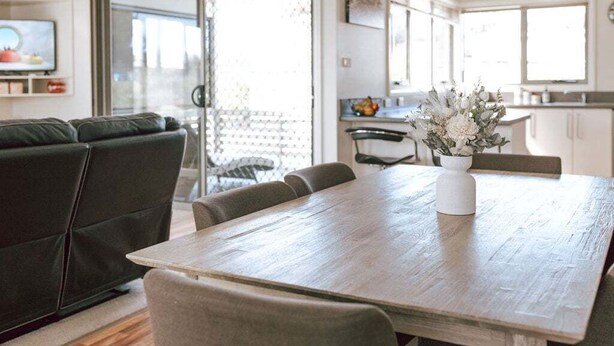 A modern living room with a wooden dining table, a green sofa, and a kitchen in the background. - Mara House - Space for Everyone (Break O'Day)