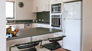 A modern kitchen with white cabinets, a black countertop, a microwave, an oven, and a refrigerator. There is a clock on the wall and a fruit bowl on the counter. - Mara House - Space for Everyone (Break O'Day)