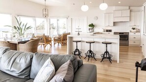 Una cocina moderna con armarios blancos, una isla central con taburetes y una zona de comedor con mobiliario de mimbre.