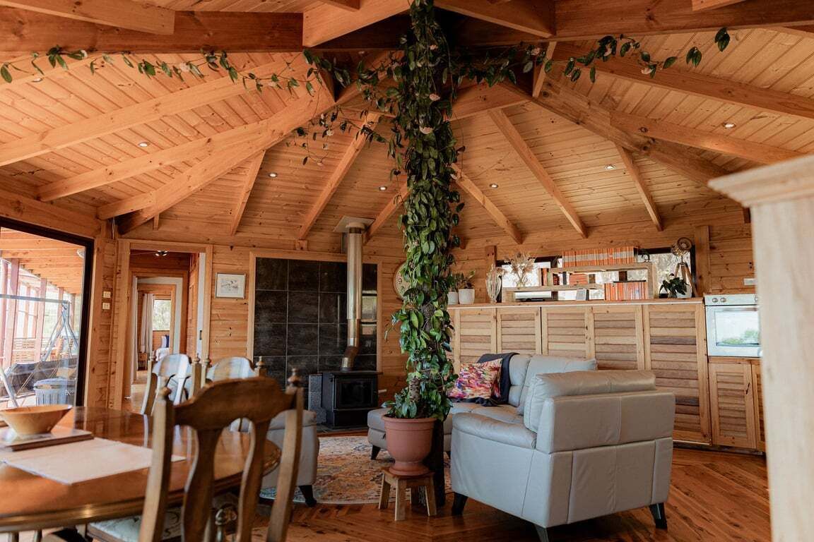 A rustic cabin interior with wooden beams, a fireplace, a dining area, and a living room with a sofa and a potted plant.