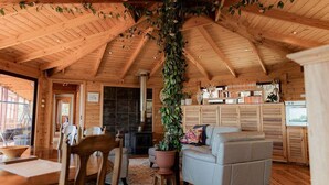 A rustic cabin interior with wooden beams, a fireplace, a dining area, and a living room with a sofa and a potted plant.