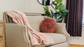 A beige sofa with a pink throw blanket and a fluffy red pillow, a green plant, and a dark curtain. - Suite Magnolia in Highland Park, Walk to Rojo (Birmingham)