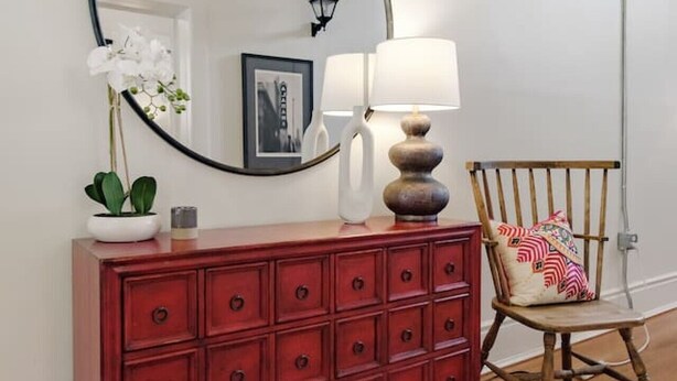 A room with a wooden floor, a red dresser with multiple drawers, a round mirror, a lamp, a chair with a cushion, and a framed picture on the wall.