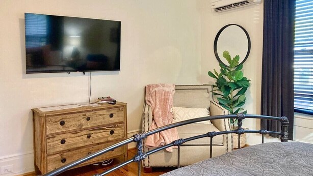 A bedroom with a bed, a wooden dresser, a television, a potted plant, and a window with curtains. - Suite Magnolia in Highland Park, Walk to Rojo (Birmingham)