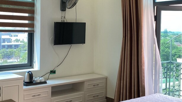A bedroom with a bed, a TV mounted on the wall, a desk with a chair, and a window with curtains. - Royal Hotel Cua Lo Beach (Cua Lo)