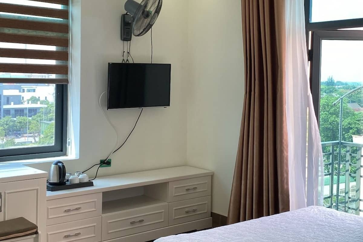 A bedroom with a bed, a TV mounted on the wall, a desk with a chair, and a window with curtains.