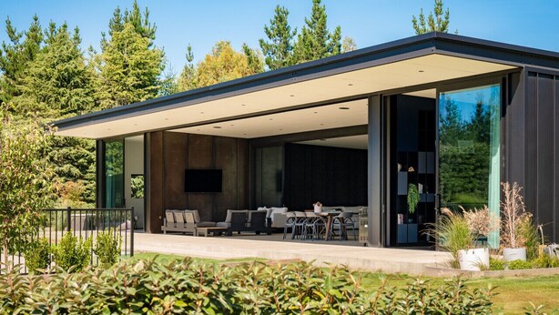 A modern house with a covered patio, large windows, and a view of a forest. - Riverbank  - Hosted by NZSIR Luxury Rental Homes (Wānaka)