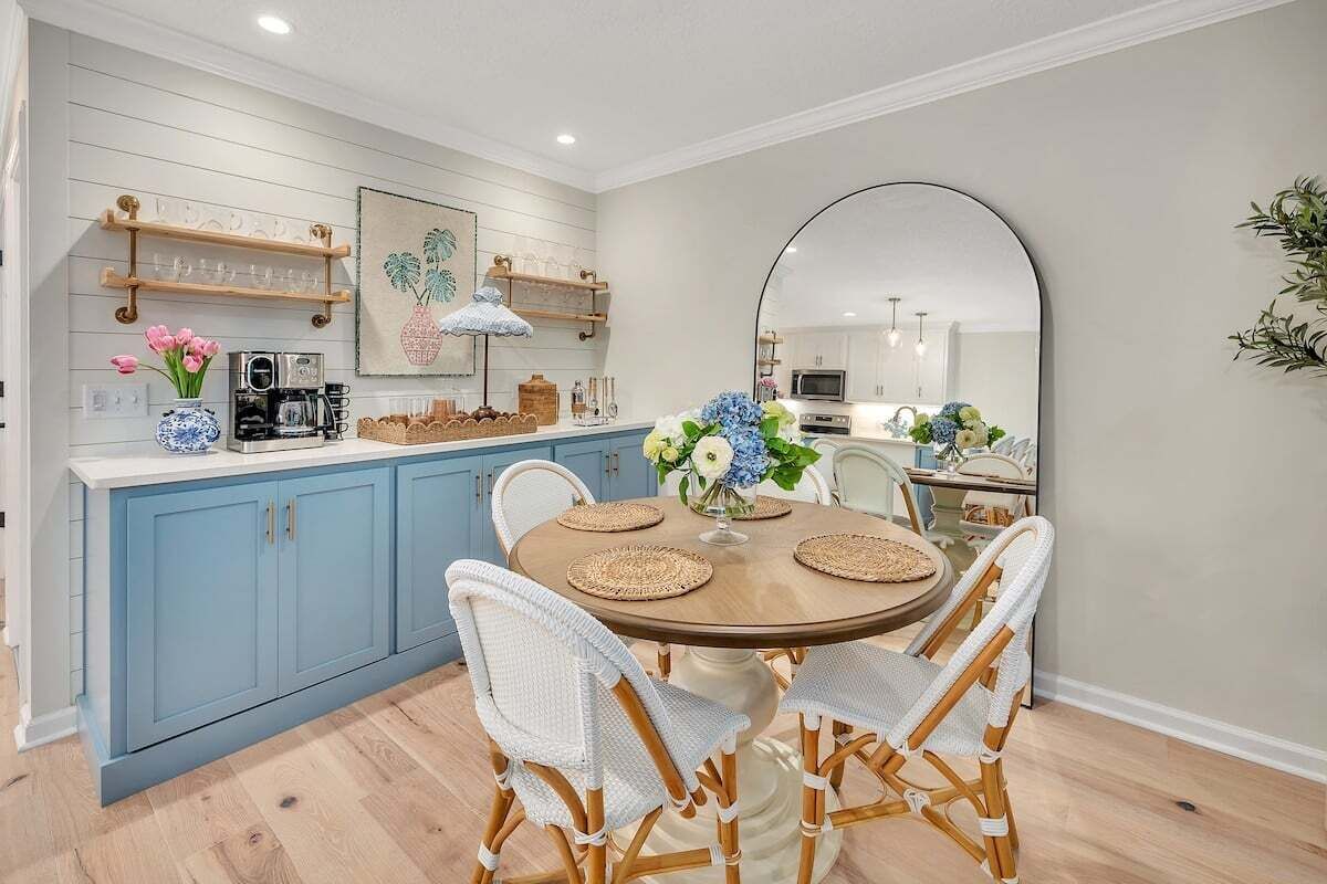 Een keuken met blauwe kastjes, een ronde houten tafel met vier stoelen en een grote spiegel aan de muur.