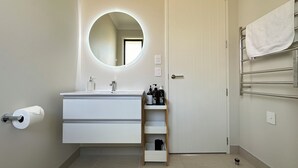 A bathroom with a white vanity, a round mirror, a towel rack, and a shelf with toiletries.