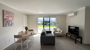 A modern living room with a dining area, a grey sofa, a TV, and a large window with a view of houses.