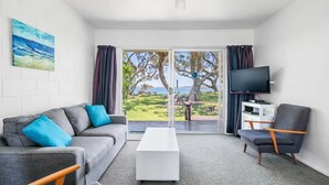 A modern living room with a grey sofa, a white coffee table, and a grey armchair. A flat-screen TV is mounted on the wall. - The Sands Motel (Tutukaka)