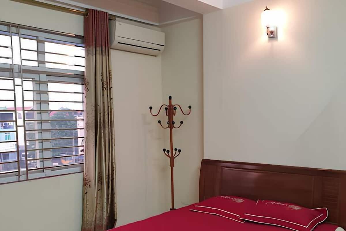 Una cama bien hecha con colcha roja y almohadas blancas, un cabecero de madera, una lámpara de pie y una ventana con cortinas.