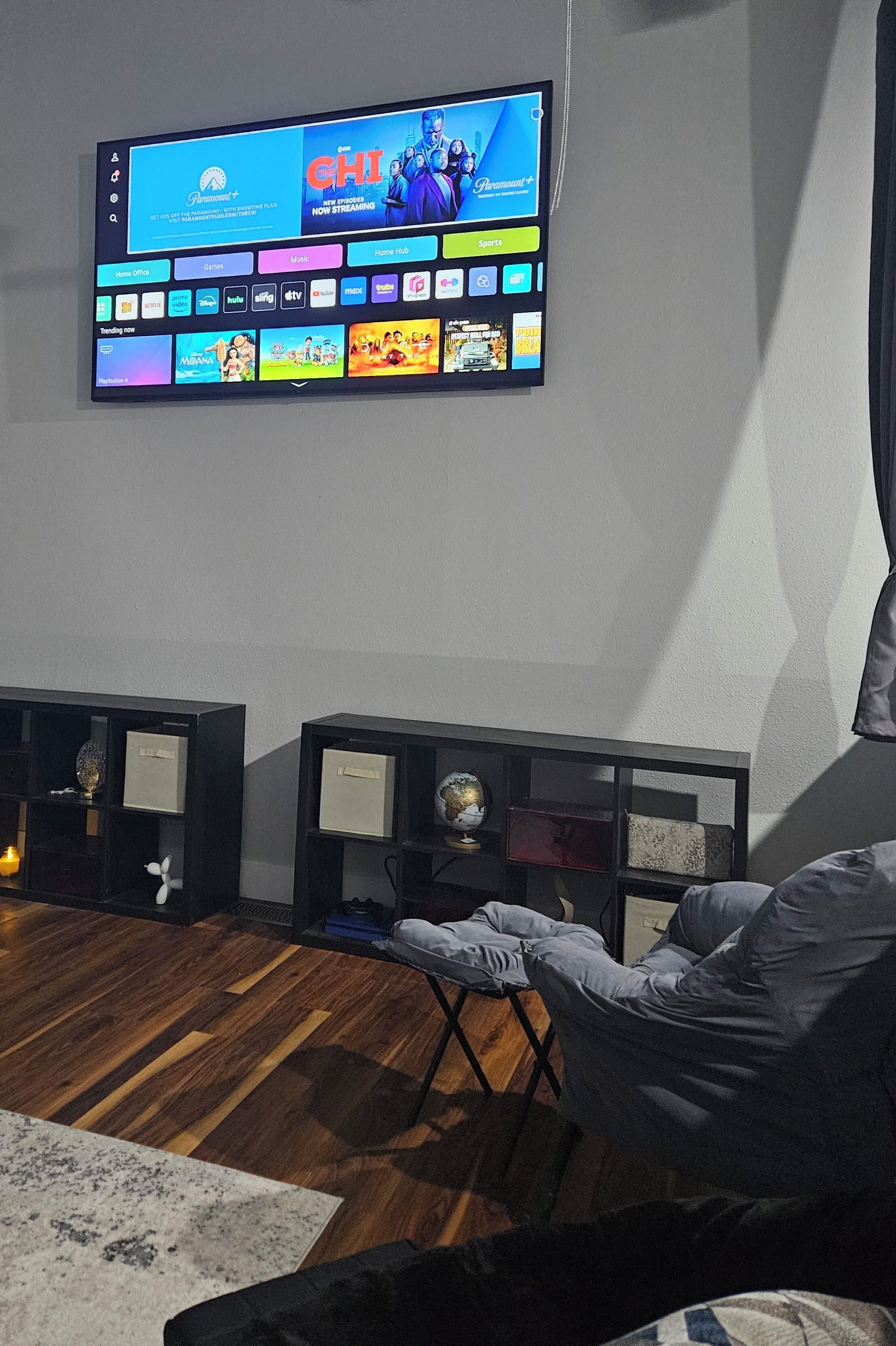 A flat-screen TV mounted on a wall displaying a streaming service interface. There are two black shelves below the TV, each containing decorative items and a small globe. A grey armchair is positioned in front of the TV.