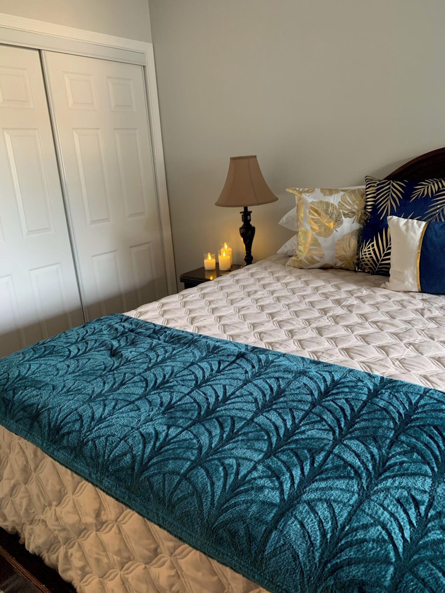 Un lit bien fait, recouvert d’une couvre-lit à motifs, une lampe de chevet et deux bougies allumées sur une table de chevet.