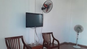 A room with a flat-screen TV mounted on the wall, two wooden chairs, a small table, and two standing fans.