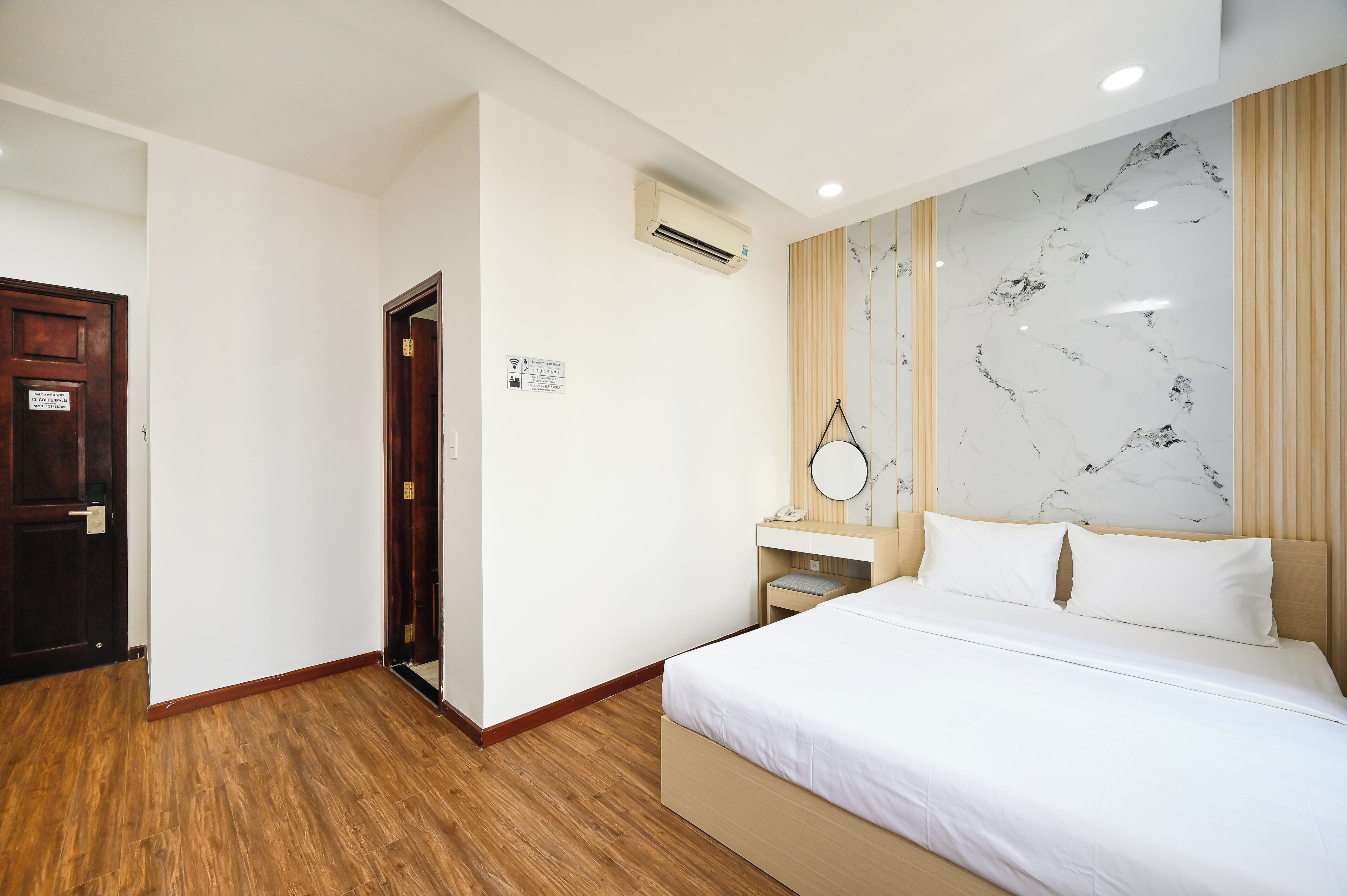 A hotel room with a bed, a wooden floor, a marble wall, a wooden door, and an air conditioning unit.