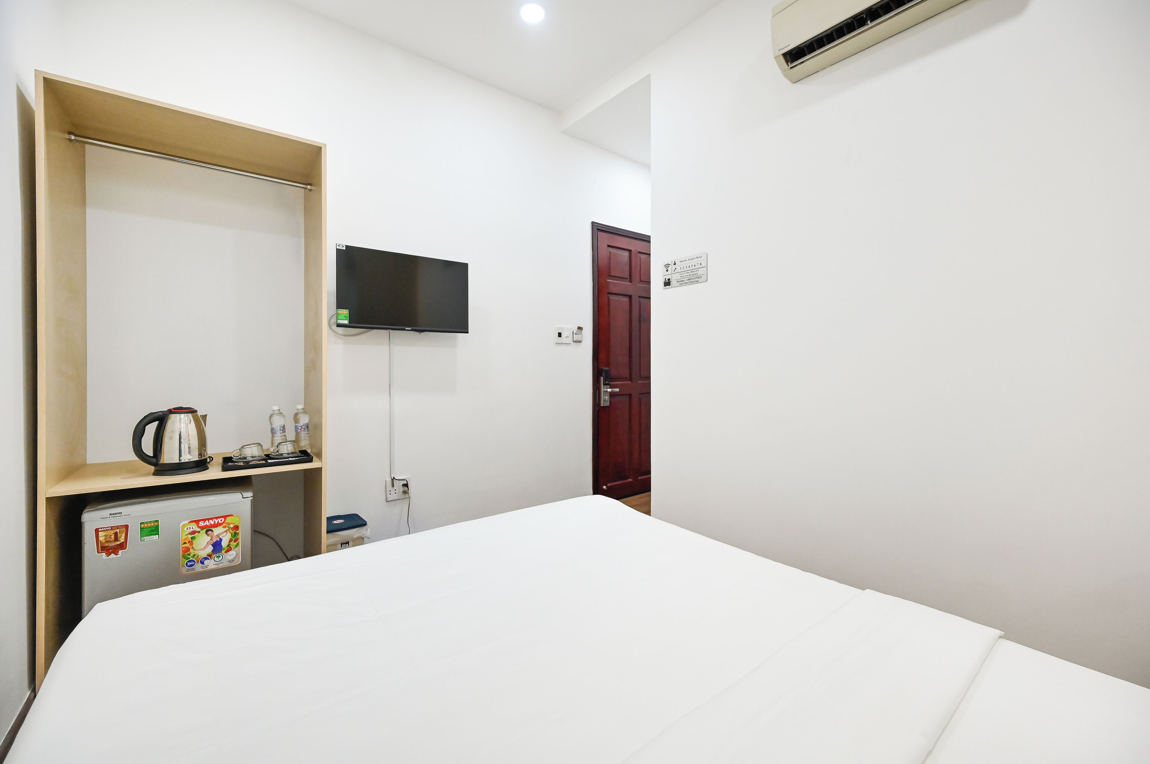 A hotel room with a bed, a TV mounted on the wall, a small fridge, a wooden shelf with a kettle, and a door with a sign.