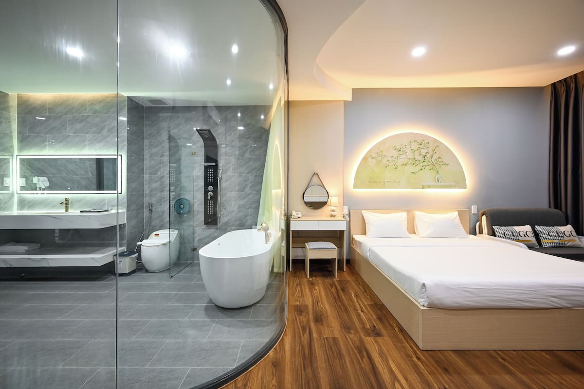 Une chambre d’hôtel moderne dotée d’une douche en verre, d’une grande baignoire, d’un plancher en bois, d’un lit avec du linge de maison blanc et d’un bureau avec une chaise.