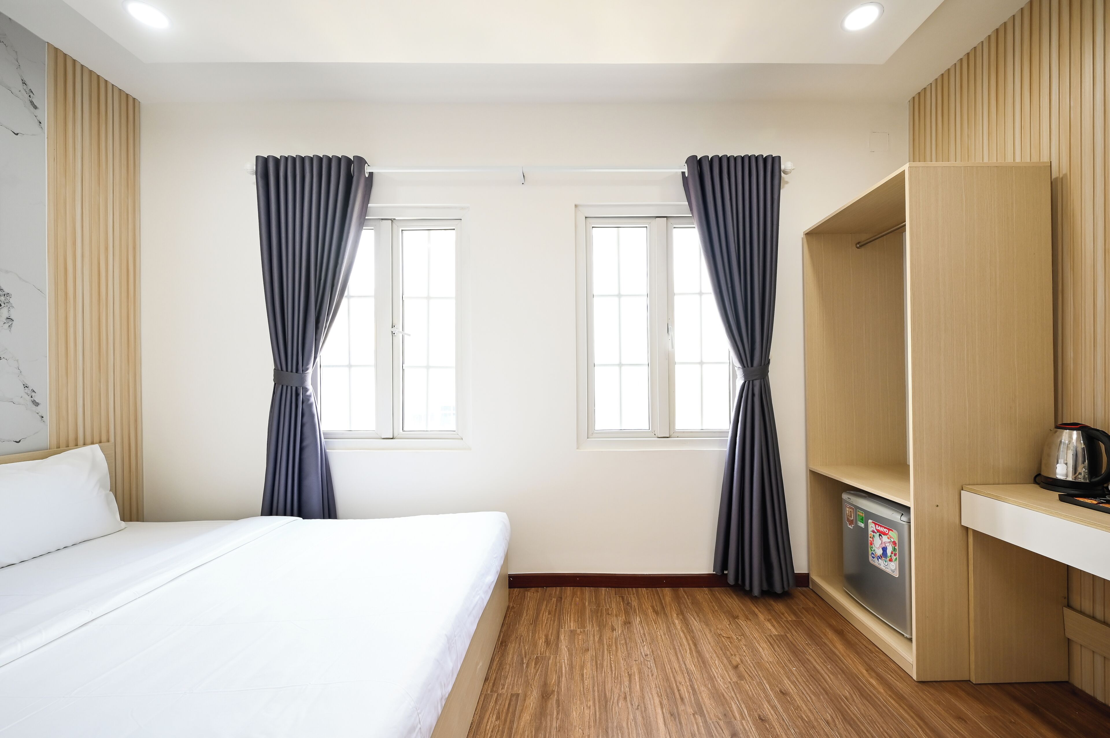 Una habitación de hotel con cama, dos ventanas con cortinas, piso de madera, estante empotrado con hervidor y refrigerador.