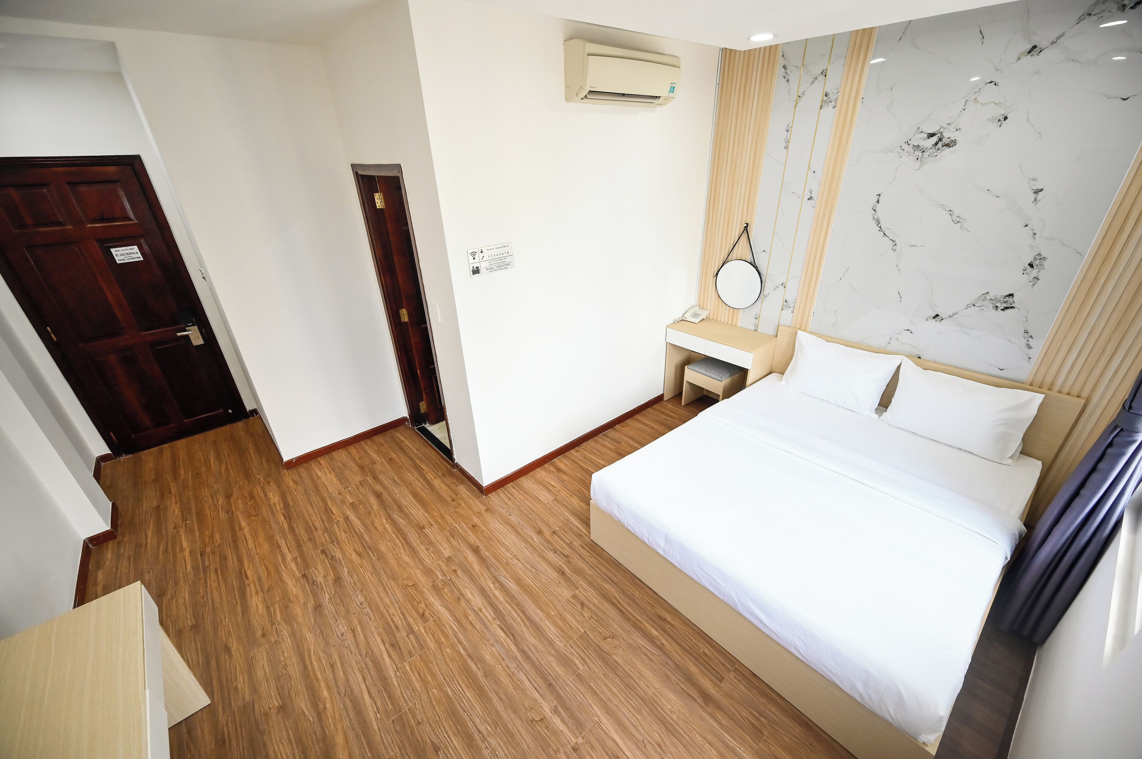 Habitación de hotel con piso de madera, cama con ropa de cama blanca, mesita de noche con lámpara y pared con diseño de mármol.