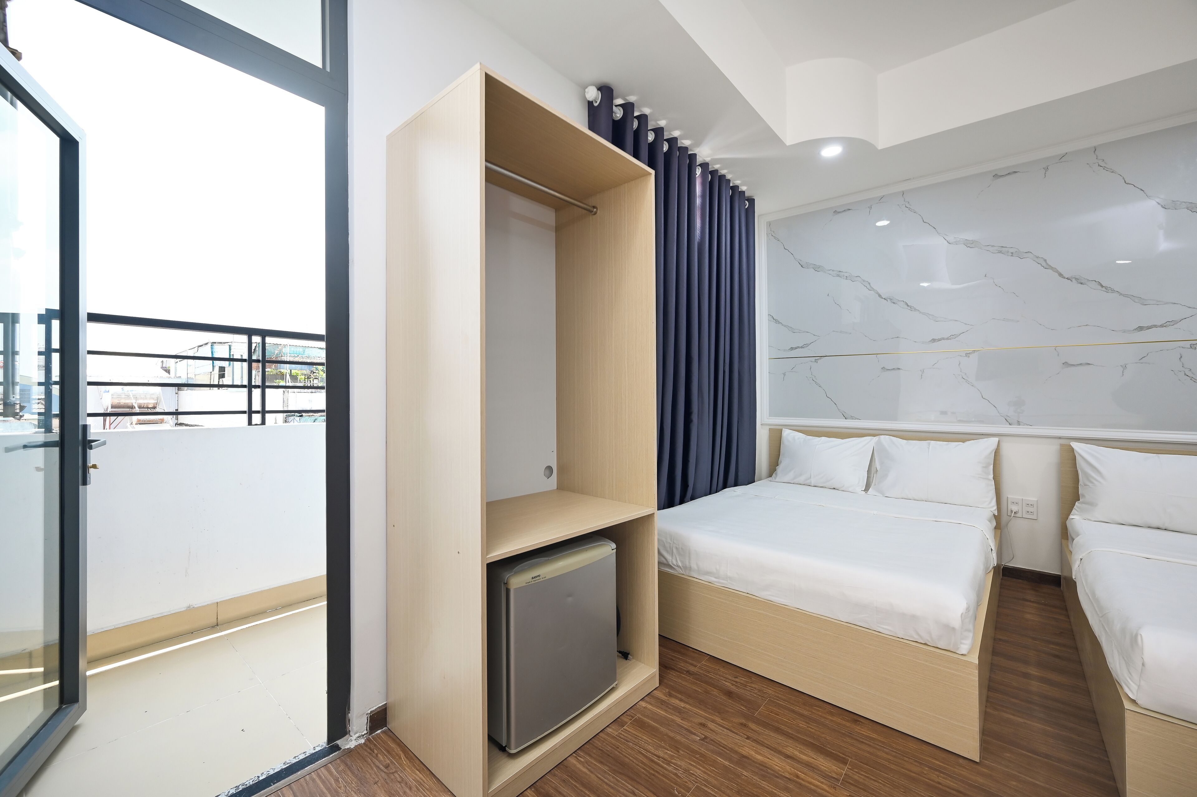 A modern hotel room with a bed, a small refrigerator, a wardrobe, and a balcony with a glass door.