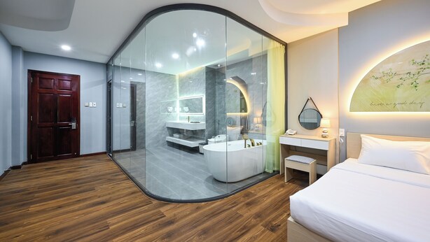 A modern hotel room with a glass partition separating the bedroom from the bathroom. The room features a wooden floor, a white bed, a small bedside table, and a wall-mounted mirror. - Sunrise Airport Hotel (Ho Chi Minh City)