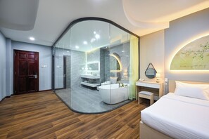 A modern hotel room with a glass partition separating the bedroom from the bathroom. The room features a wooden floor, a white bed, a small bedside table, and a wall-mounted mirror. - Sunrise Airport Hotel (Ho Chi Minh City)