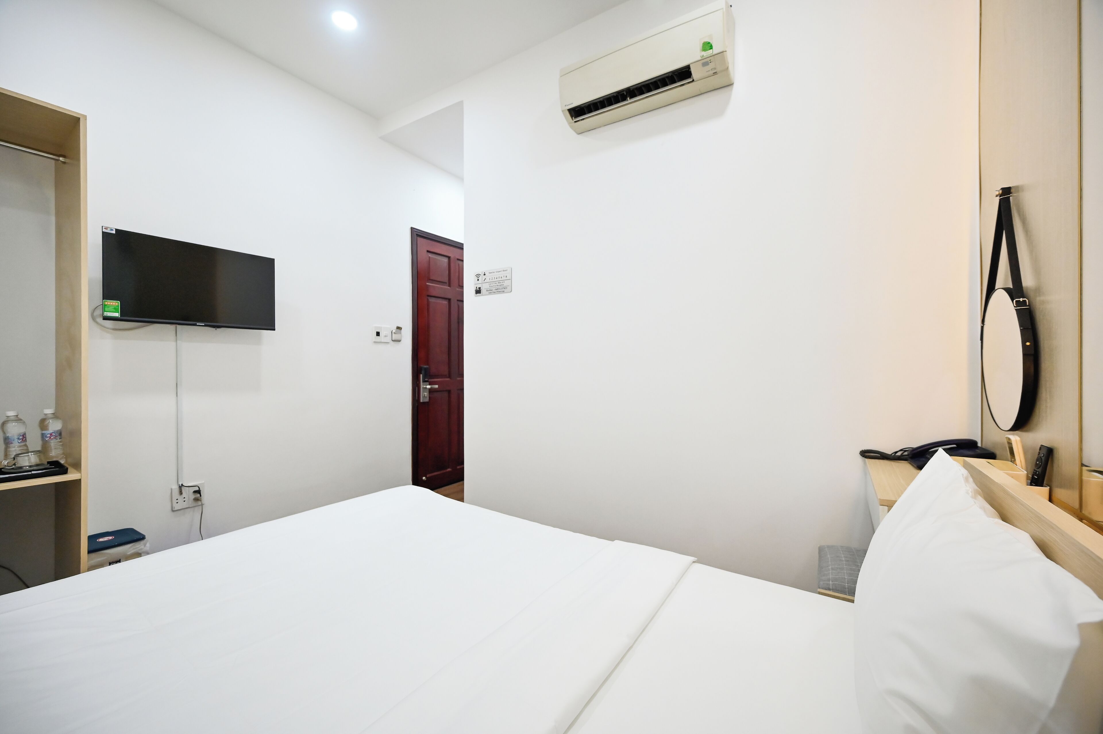 Habitación de hotel con cama, televisor montado en la pared, equipo de aire acondicionado, estante de madera con artículos de aseo y mesita de noche con teléfono y control remoto.