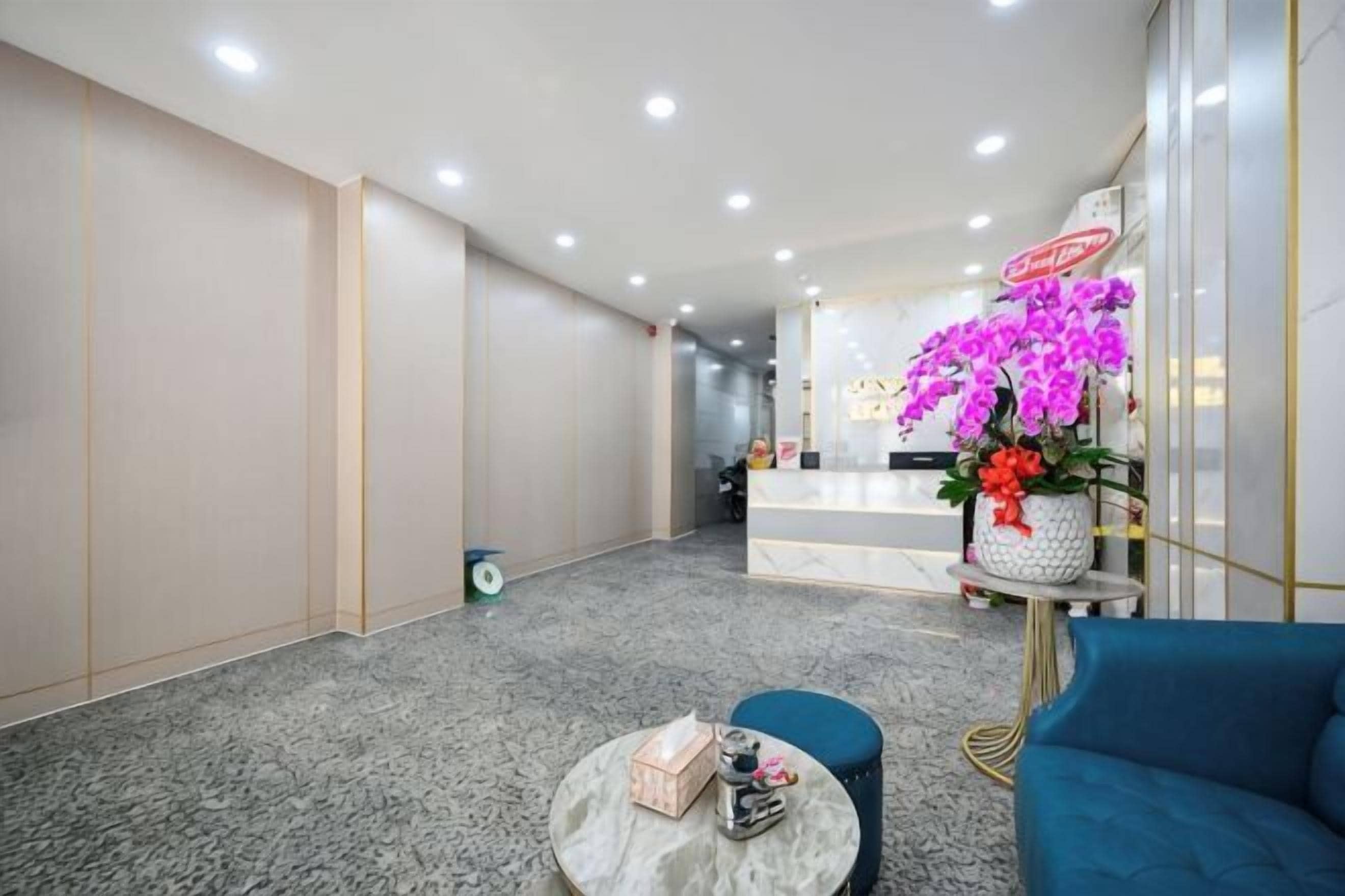A hotel lobby with a reception desk, a blue sofa, a small round table with items, and a decorative flower arrangement.