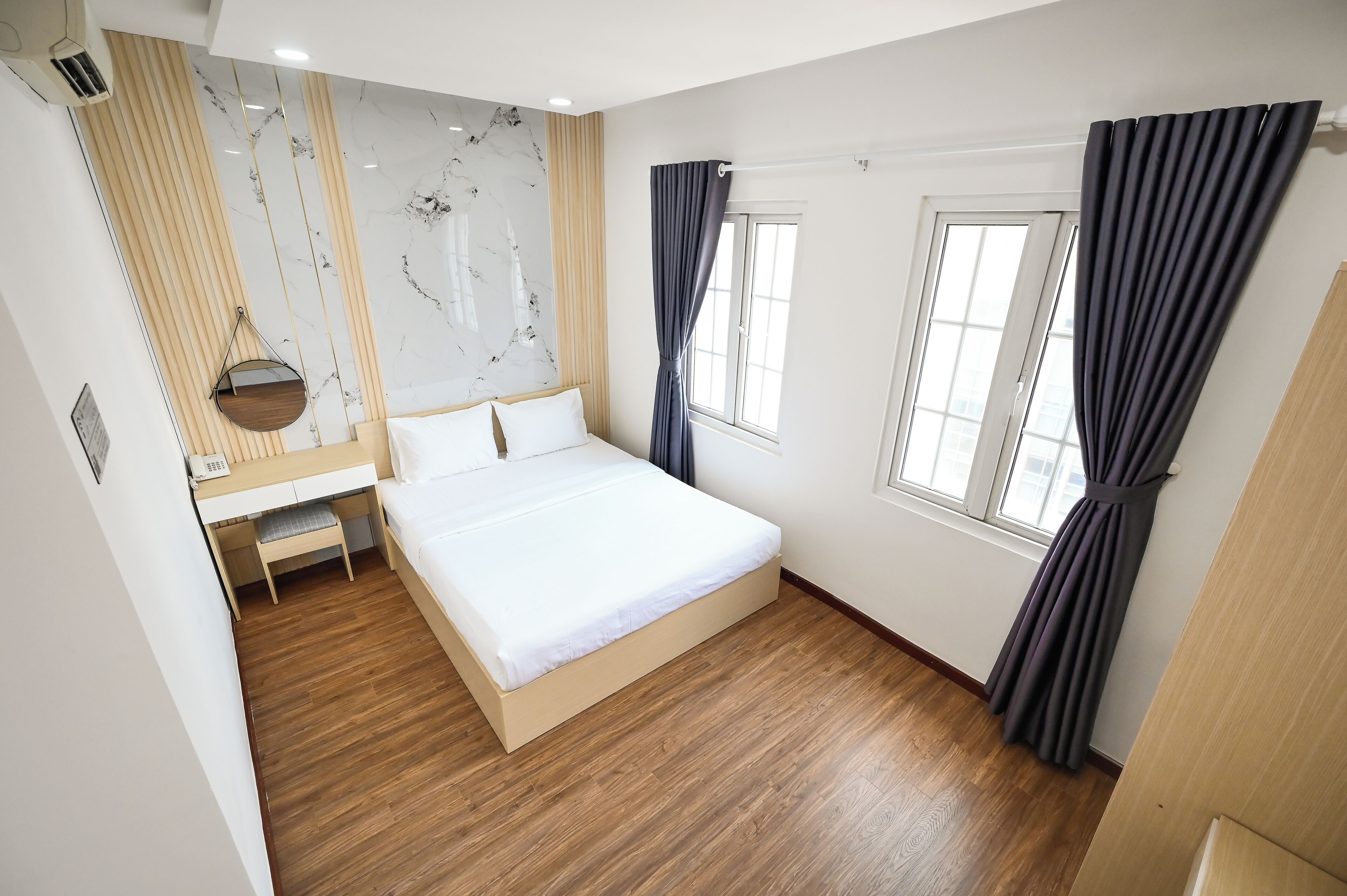 A modern hotel room with a wooden floor, a bed with white bedding, a bedside table with a lamp, and a window with curtains.
