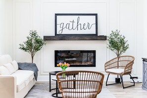 A living room with a fireplace, a sofa, two wicker chairs, a coffee table, and a framed sign above the fireplace that reads "gather".