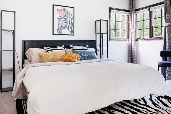 A neatly made bed with a zebra-patterned rug, a framed zebra artwork on the wall, and a window with curtains.
