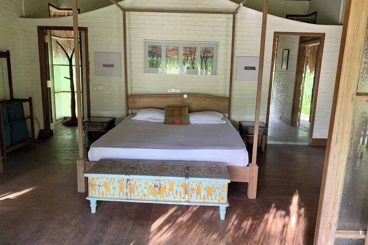 Una cama con estructura de madera y un banco frente a ella, una pared blanca decorada con cuadros, y suelo de madera.